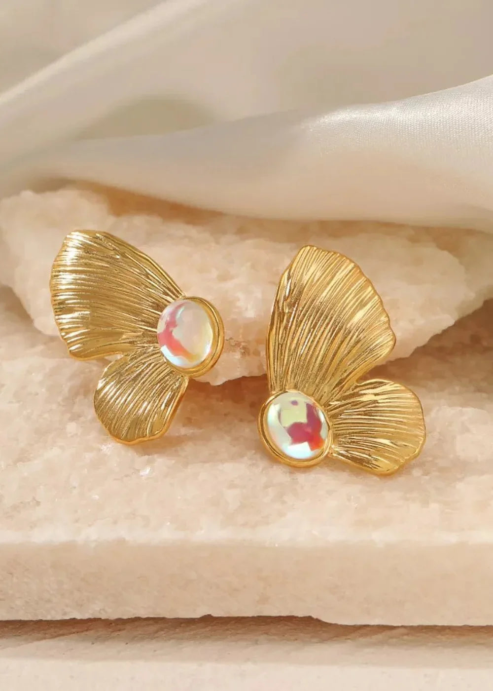 Gold-plated butterfly earrings with iridescent resin accents on a textured surface.