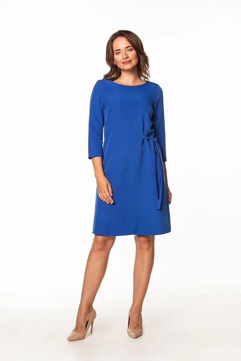 Full-length shot of a woman in a royal blue, knee-length dress with 3/4 sleeves and a tie waist, paired with nude heels.
