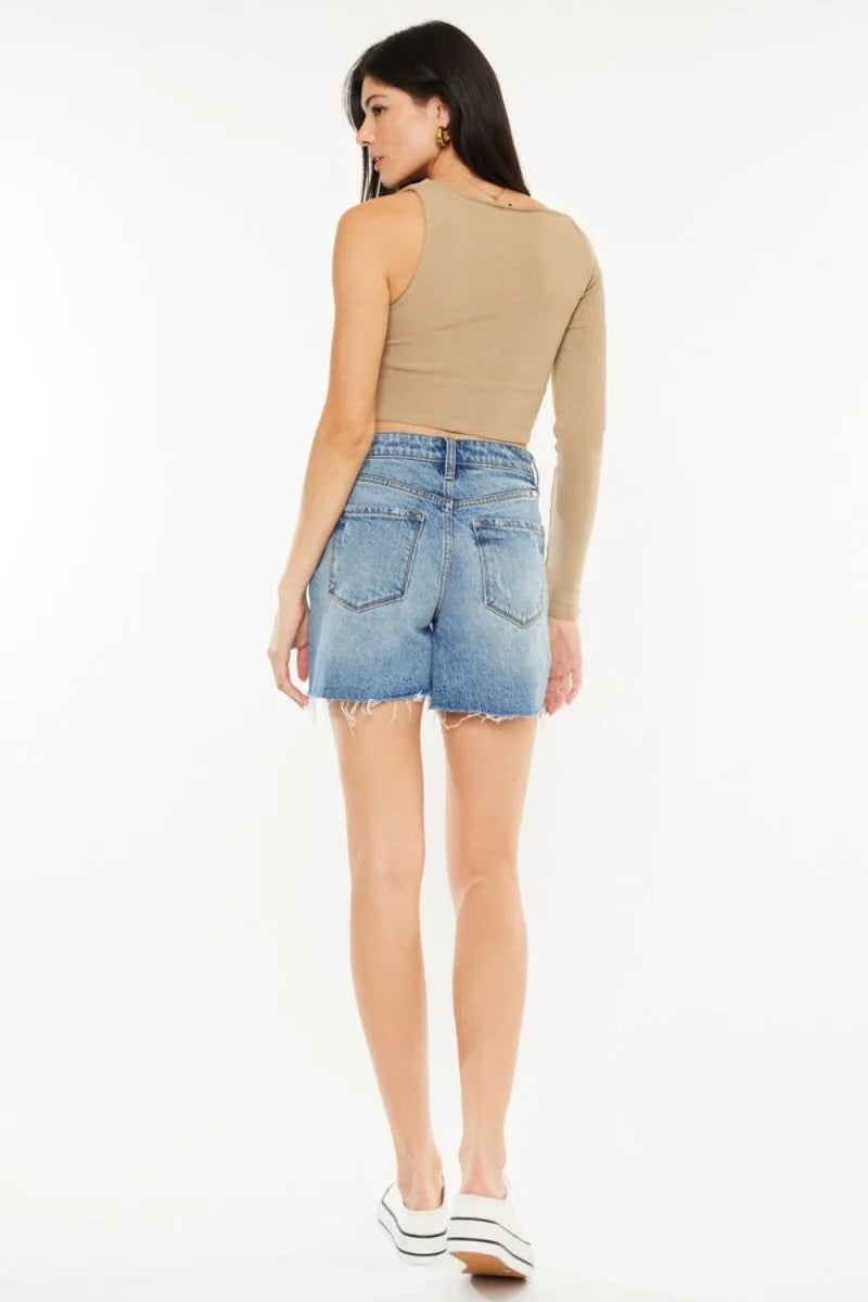 Back view of distressed denim shorts with frayed hem, on a model wearing a tan one-sleeve top.