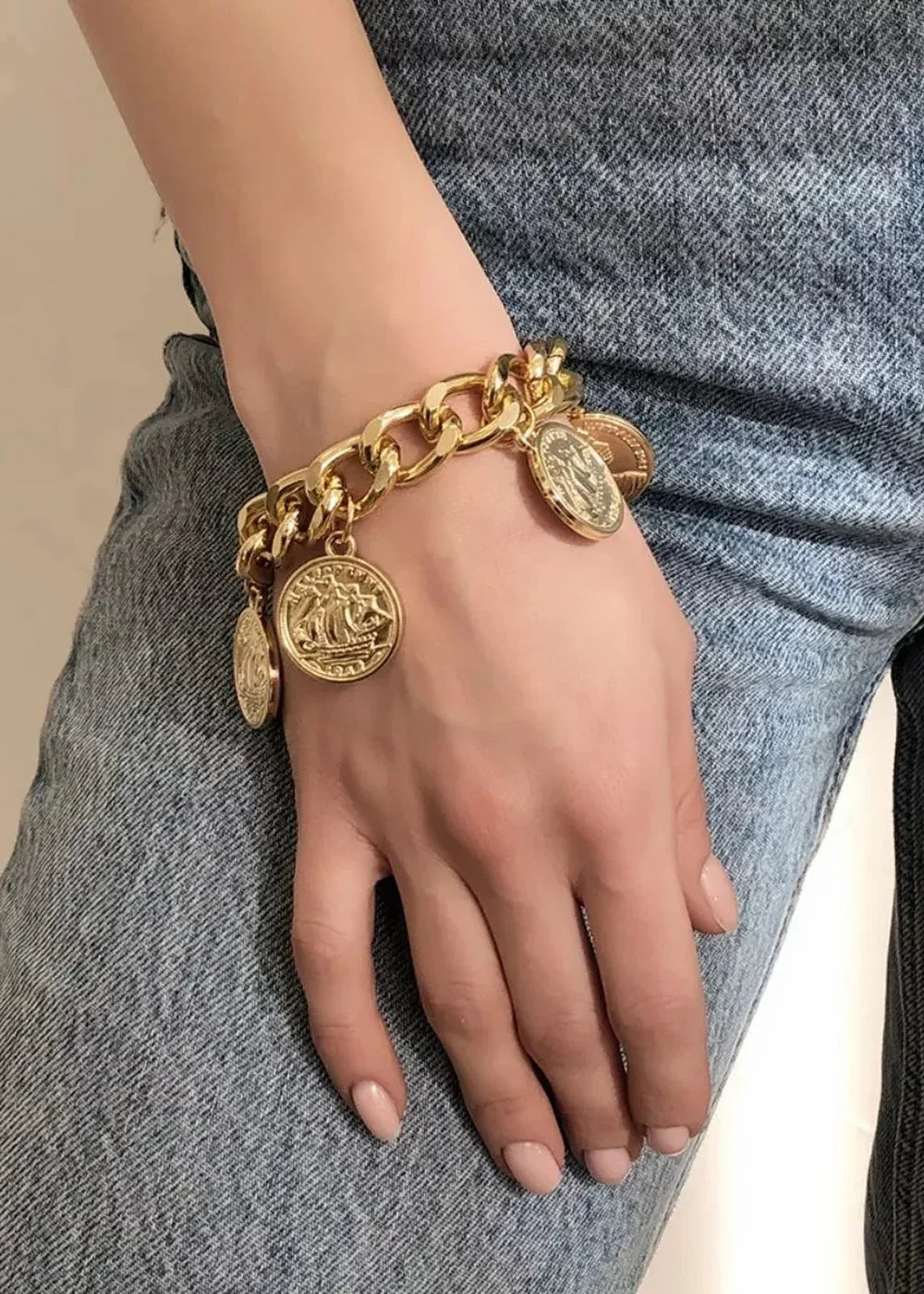Gold-plated charm bracelet with coin pendants on a hand in front of blue denim.