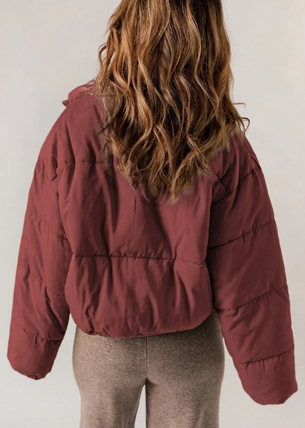 Back view of a woman wearing a cropped, brick red puffer jacket with horizontal quilting and brown knit pants.