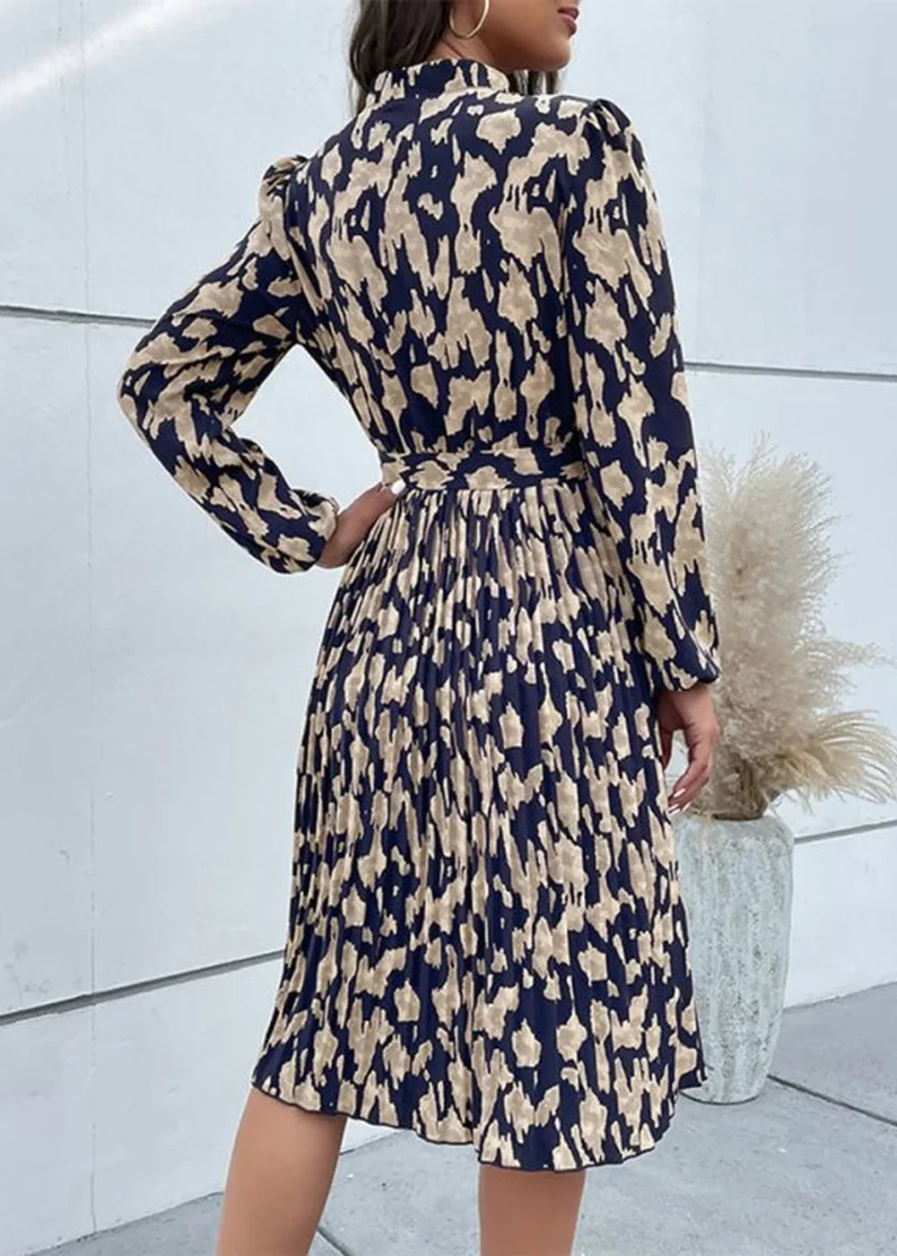 Back view of a woman wearing a navy and tan abstract print pleated midi dress with long sleeves and a belted waist.