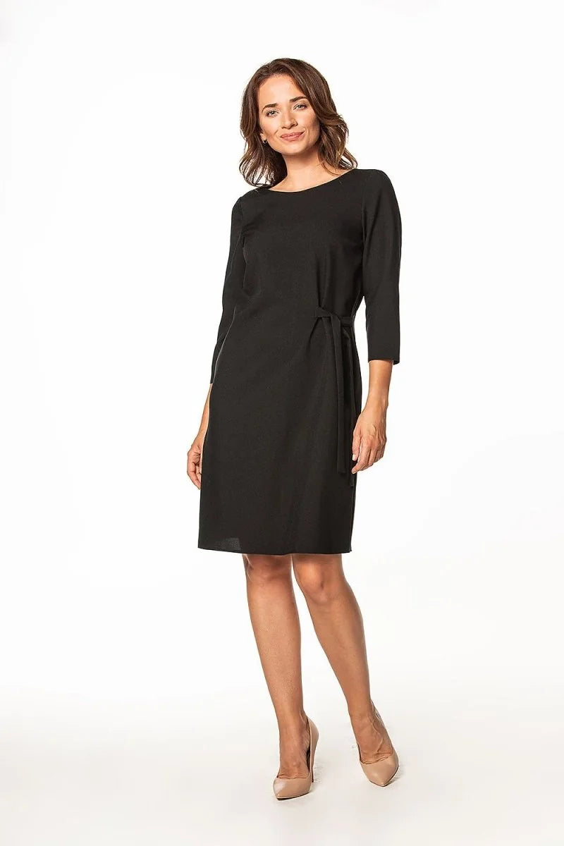 Woman in a black, knee-length dress with a tie waist and three-quarter sleeves, paired with nude pumps.