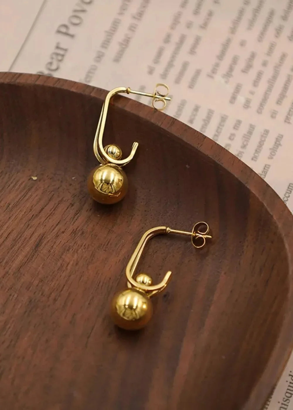 Close-up of two shiny gold hoop earrings with small gold balls hanging from them, resting on a wooden surface.