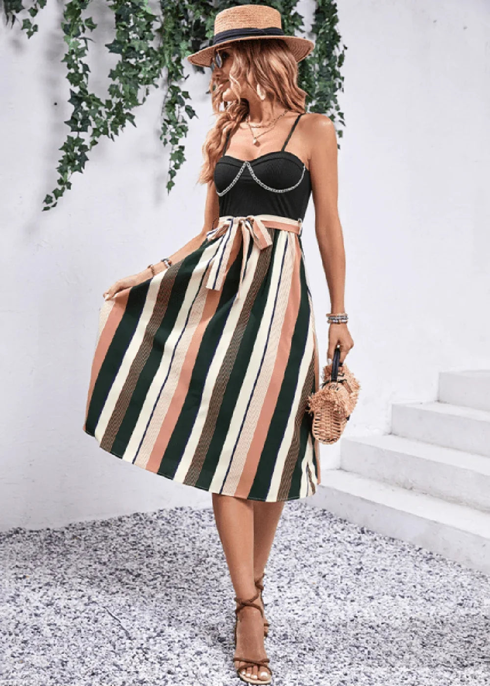 Woman in a black sweetheart neckline dress with a brown, pink, green, and white striped skirt, straw hat, and wedge sandals.