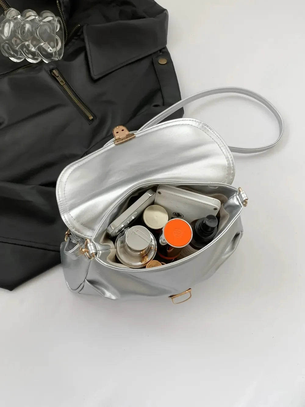 Silver metallic handbag overflowing with makeup and a phone, next to a black jacket.