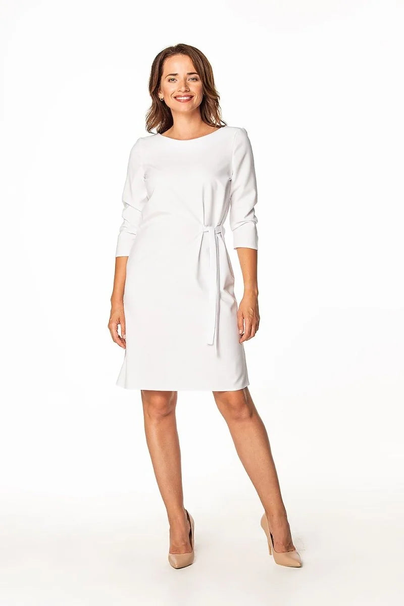 Woman in a white, knee-length sheath dress with a tie waist and three-quarter sleeves, paired with nude pumps.