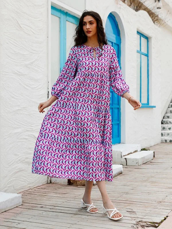 Tiered midi dress in a geometric pattern of pink, purple, and light blue, with a tie-neck and flowing sleeves.
