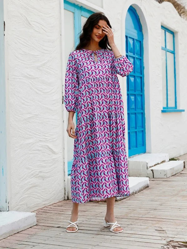Woman in tiered midi dress with geometric pattern of pink, purple, and blue shapes, wearing white sandals.