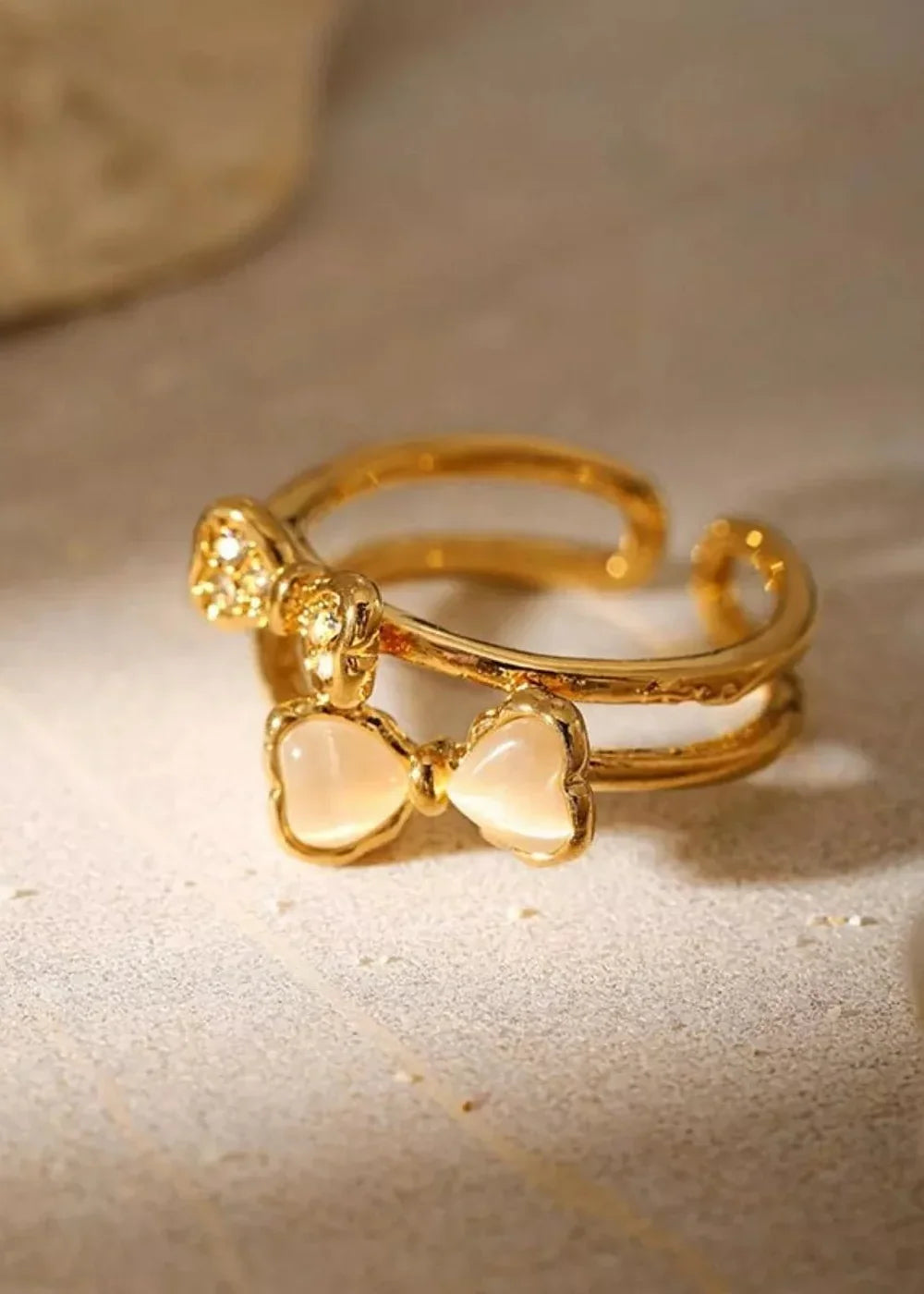 Gold double-band ring with two bow charms, one with rhinestones, the other with a milky stone.