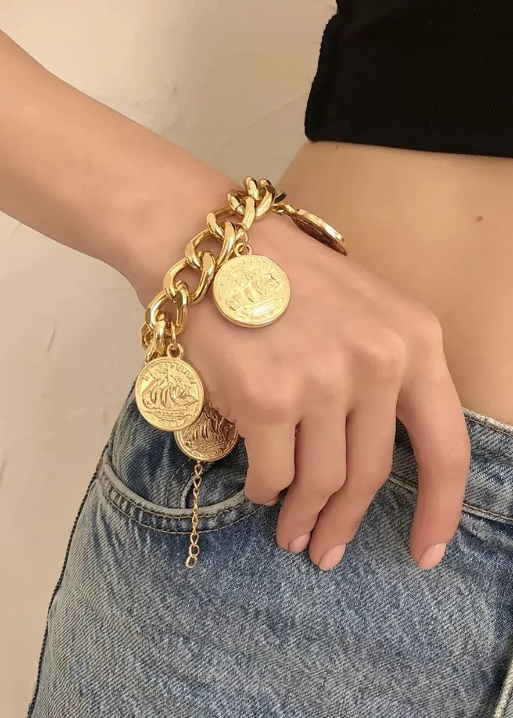 Gold coin charm bracelet with chunky chain, worn on a hand against blue denim.