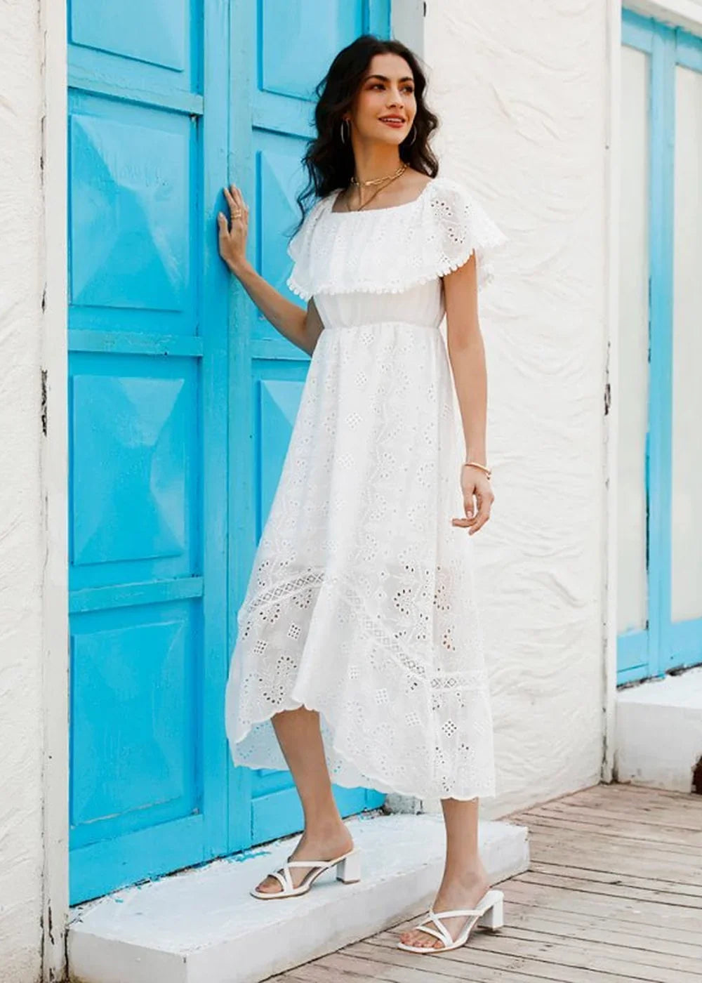 White eyelet midi dress with ruffled neckline and flowy skirt, worn by a woman standing by a blue door.