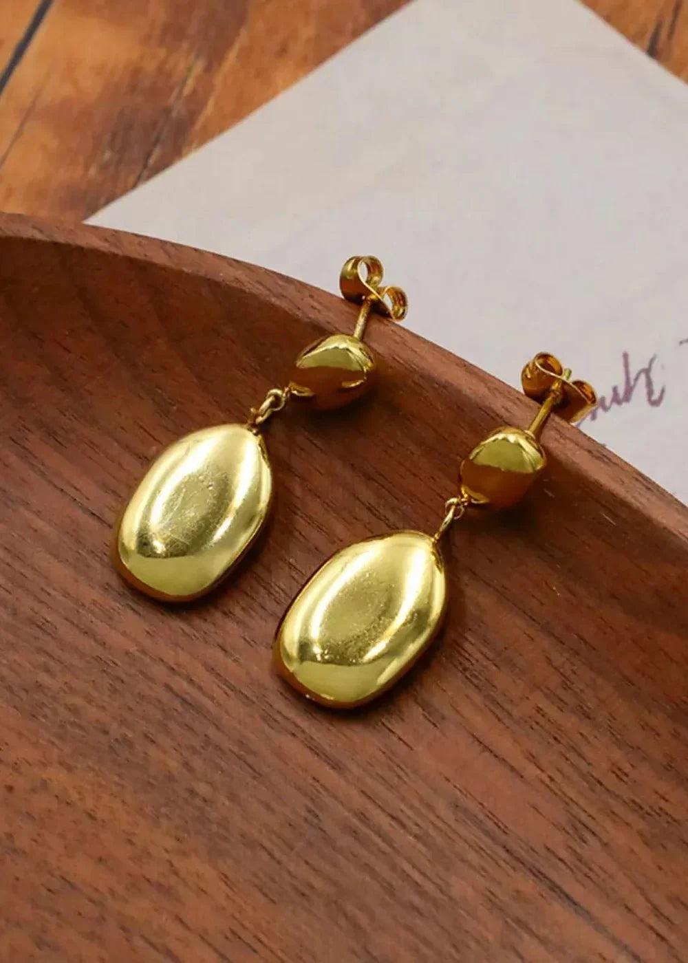 Gold oval drop earrings with textured studs, displayed on a wooden tray.