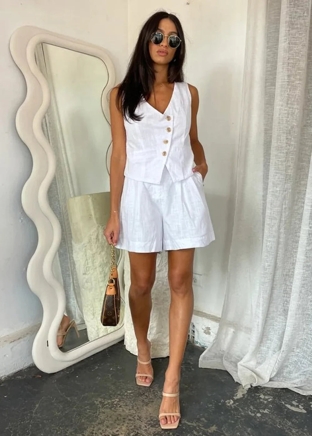Woman in white linen vest and shorts set with gold buttons, holding a brown monogram bag.