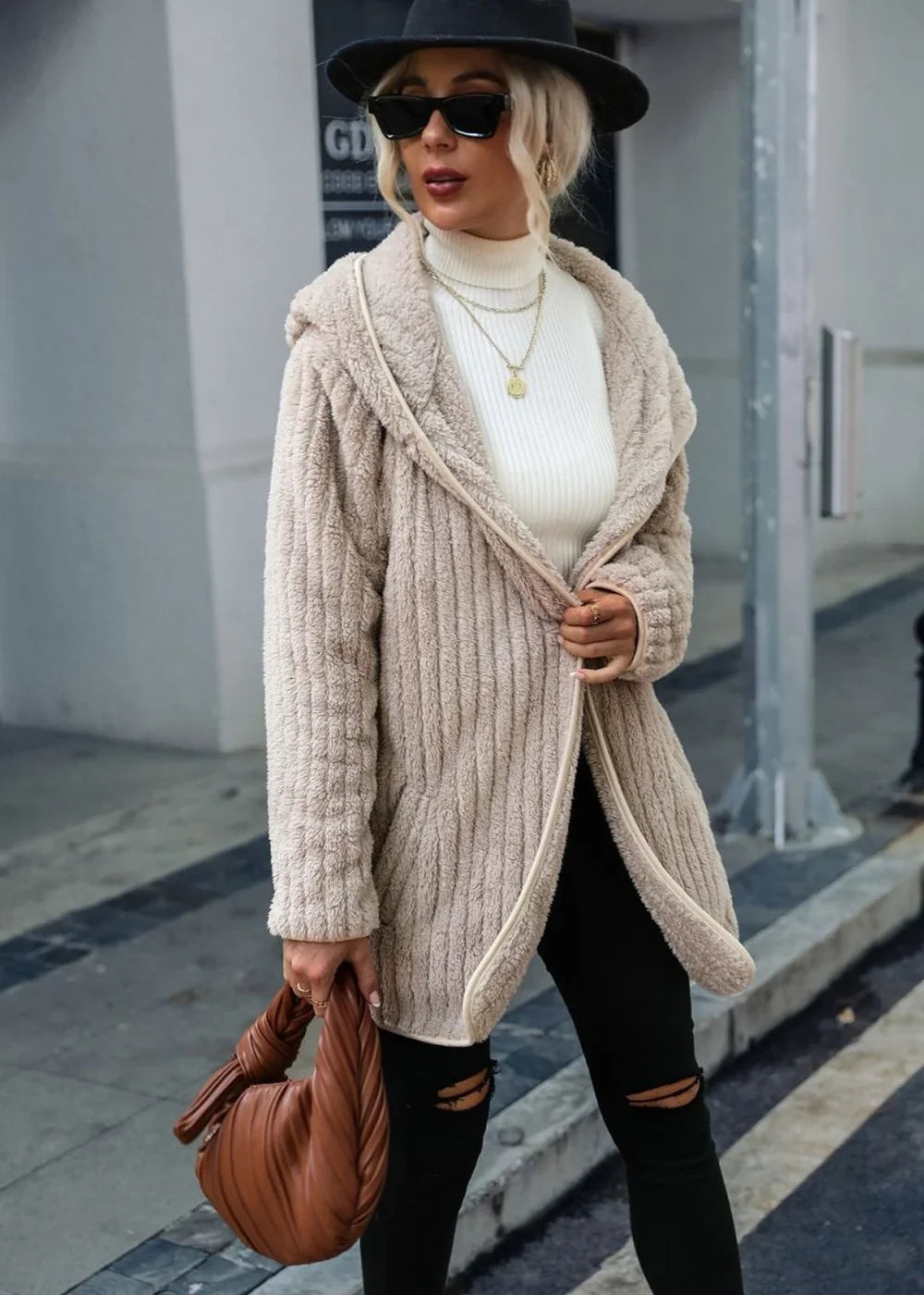 Model in a fuzzy, textured beige hooded cardigan over a white turtleneck, black ripped jeans, and a brown knotted handbag.