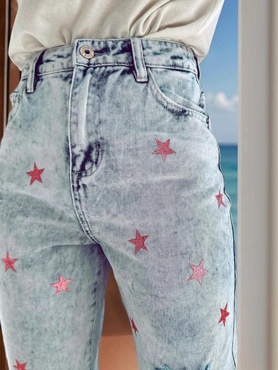 Light wash denim jeans with scattered pink glitter star pattern and frayed hem.