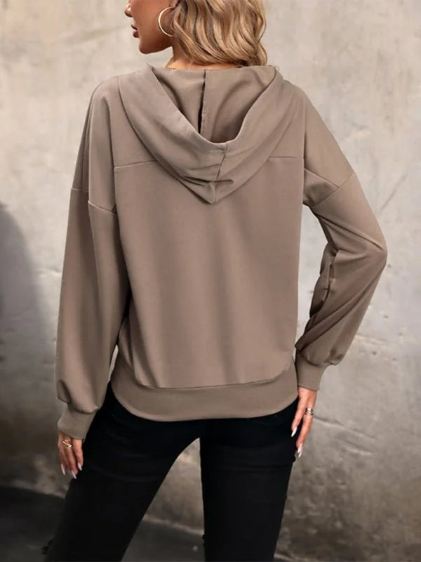 Back view of a woman wearing a taupe hooded sweatshirt with black pants and hoop earrings.