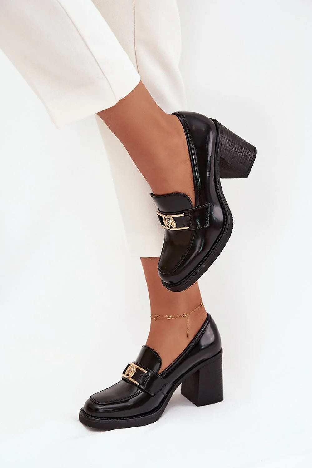 Black patent leather loafers with gold buckle detail and block heel, worn with white pants and anklet.