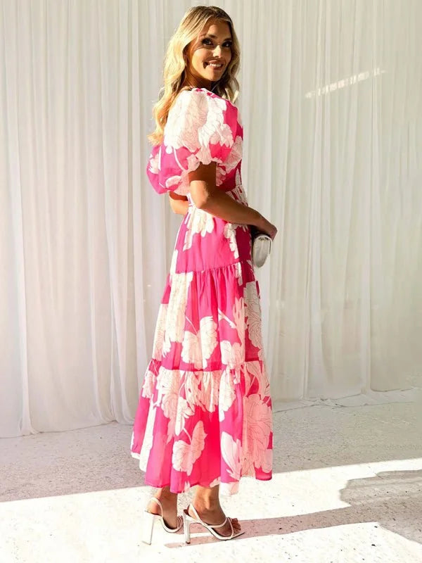 Woman in a vibrant pink tiered maxi dress with a large white floral print and puff sleeves, styled with white heeled sandals.
