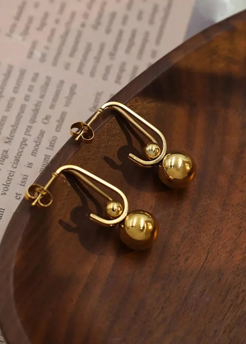 Shiny gold-plated earrings with a modern U-shaped hook and two spherical charms, resting on a wooden tray over a textured ...
