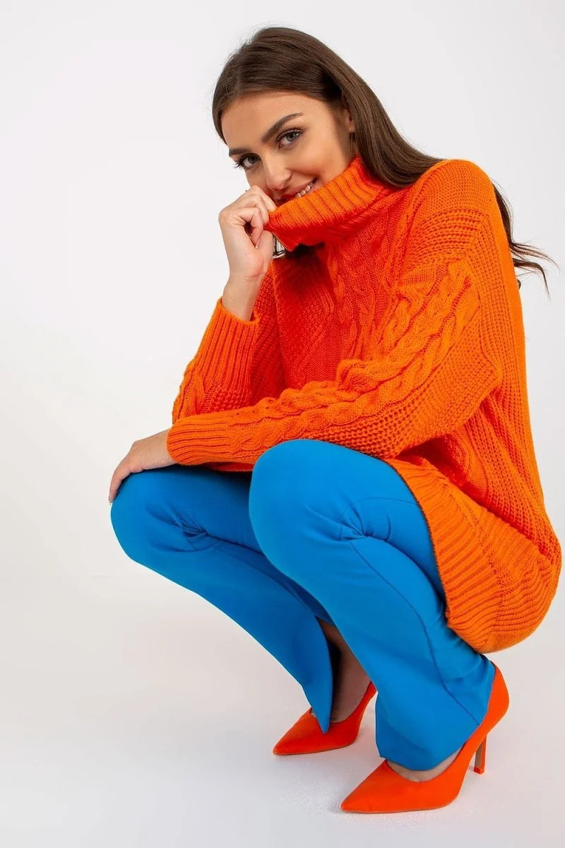 Bright orange cable-knit turtleneck sweater with blue pants and orange heels.