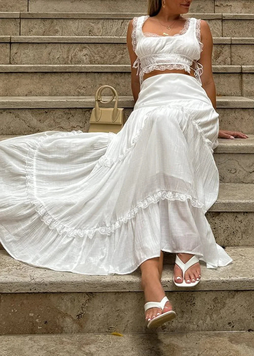 White tiered maxi skirt with lace trim and a ruffled hem, worn by a model sitting on stone steps.