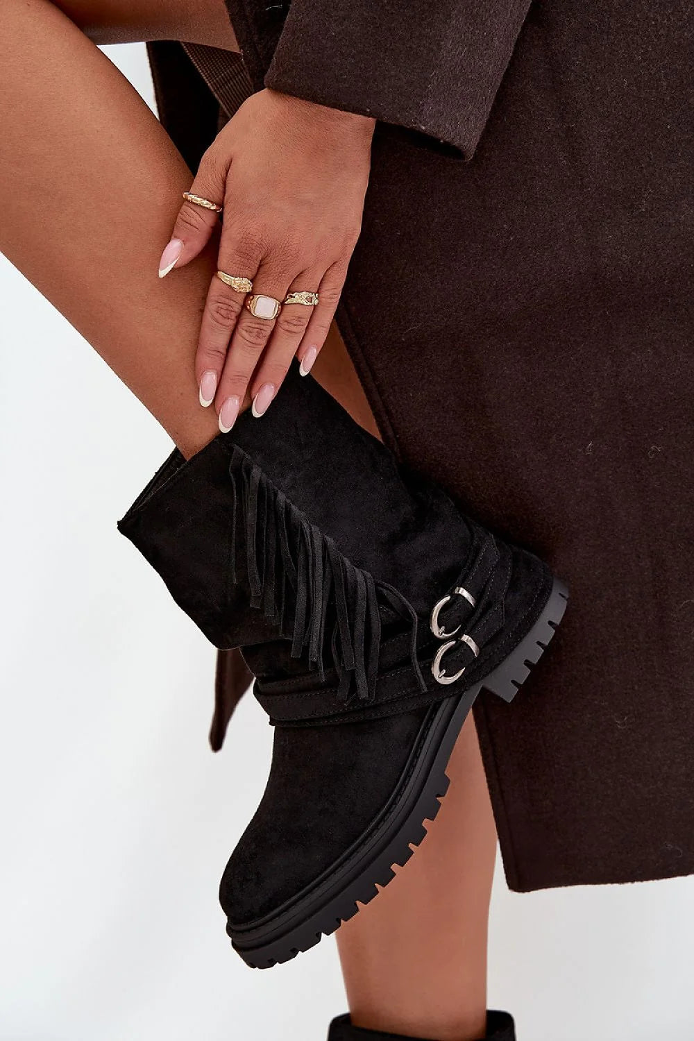 Black suede ankle boots with fringe detail and double buckle straps on a model's leg.