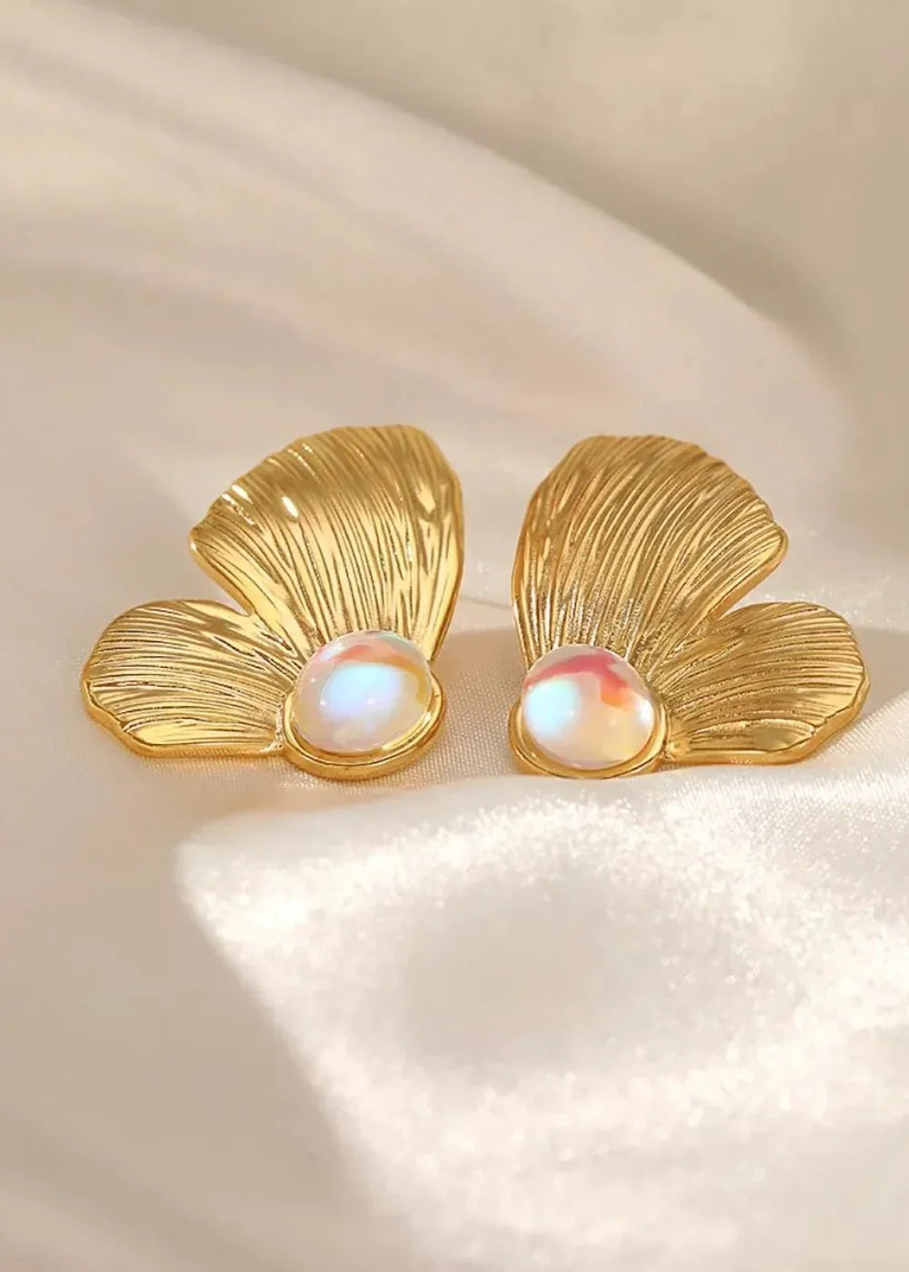 Golden, fan-shaped earrings with iridescent, pearlescent stones, set against a soft, draped ivory fabric.