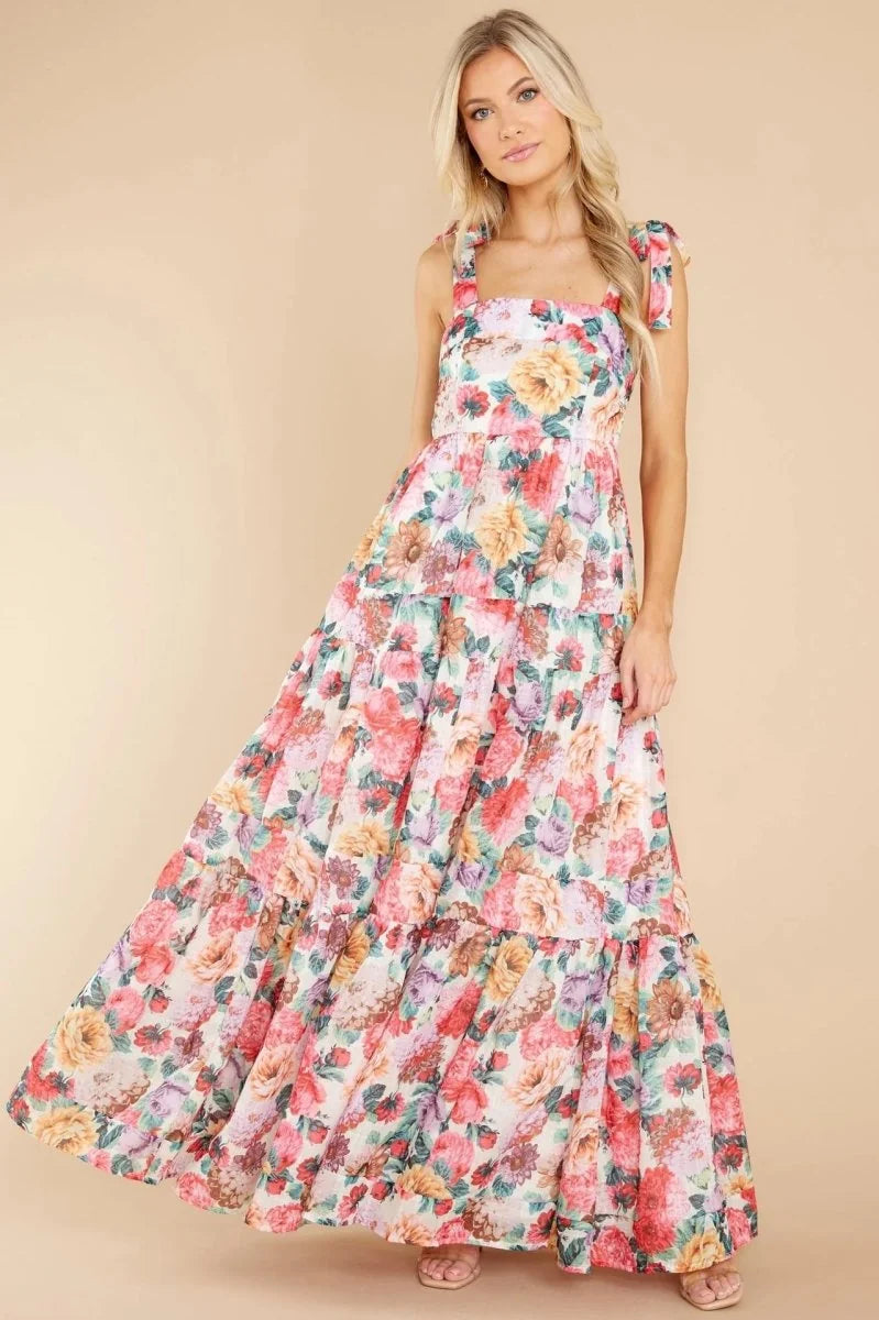 A woman in a flowing, tiered floral maxi dress with tie straps stands against a neutral background.