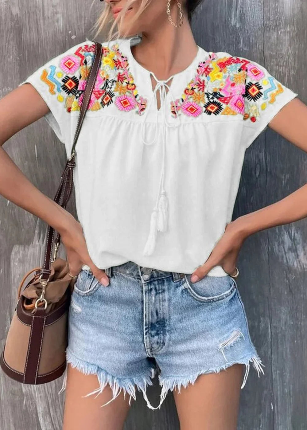 White peasant top with colorful embroidered yoke and tassels, paired with distressed denim shorts.