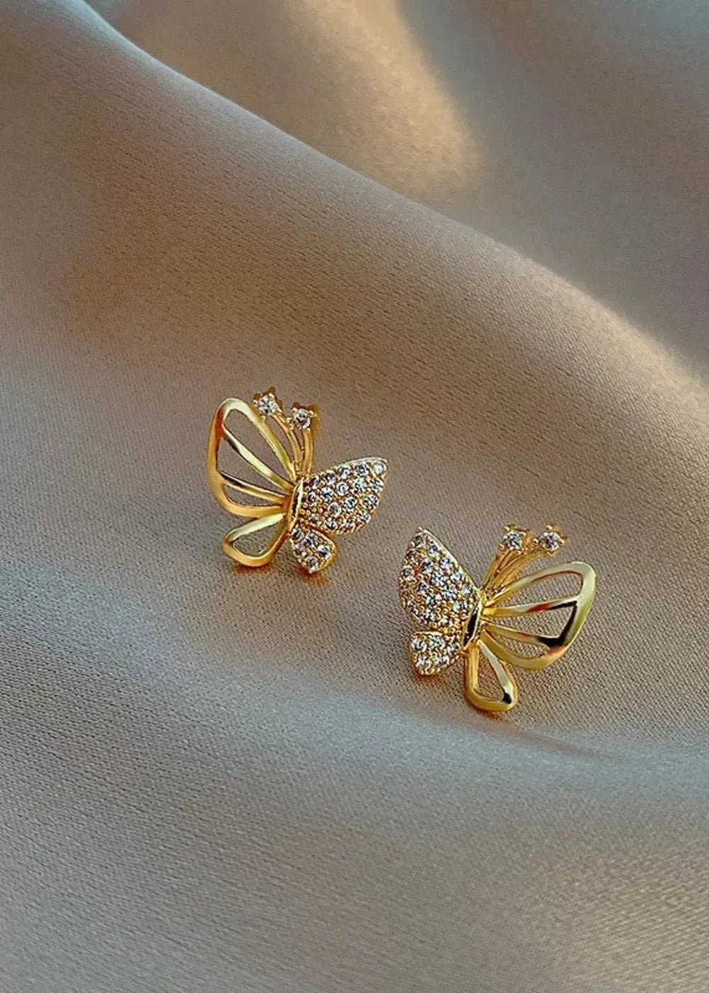 Gold butterfly earrings with rhinestones against a textured beige fabric background.