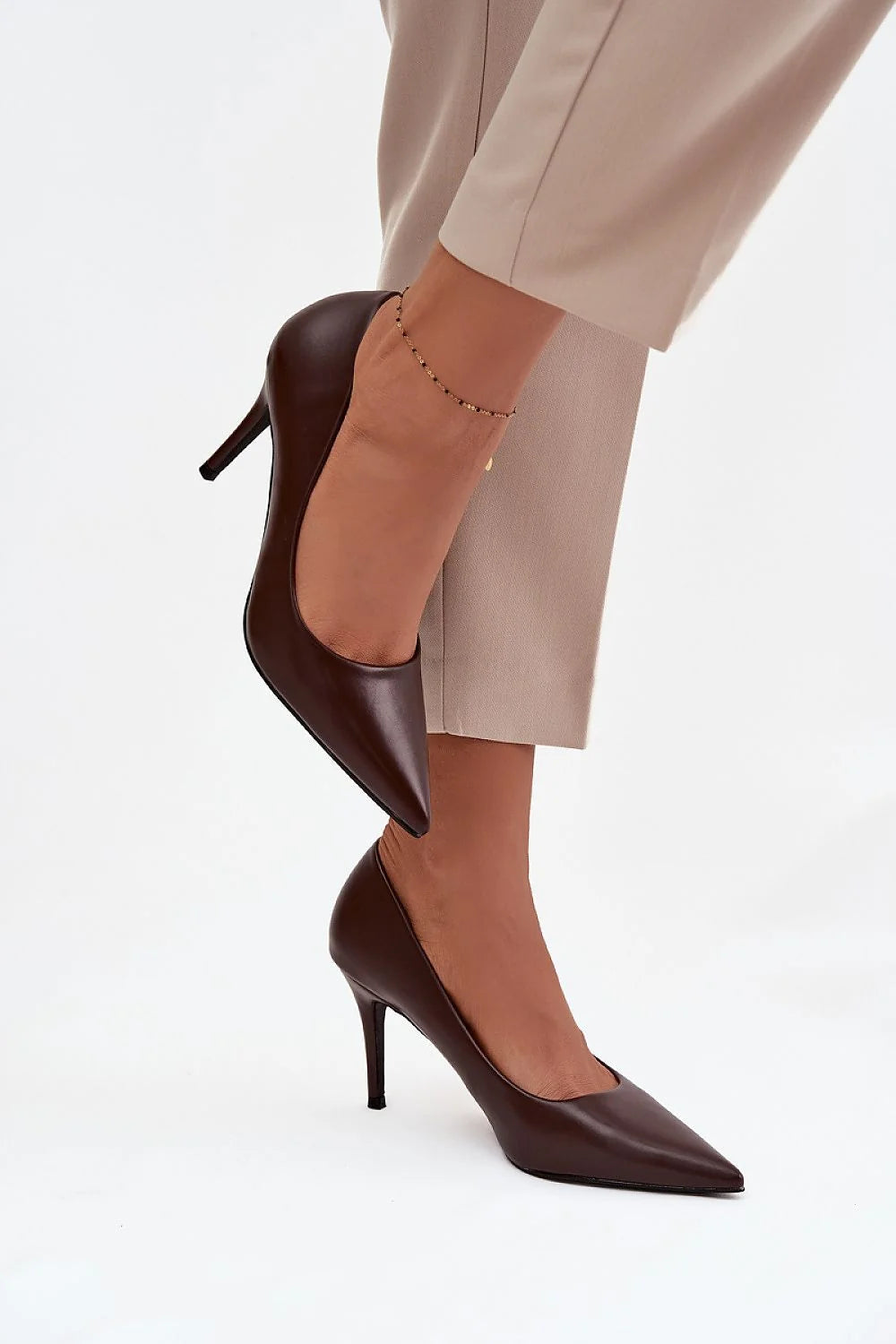 Brown pointed-toe pumps with stiletto heels, worn with a gold anklet.