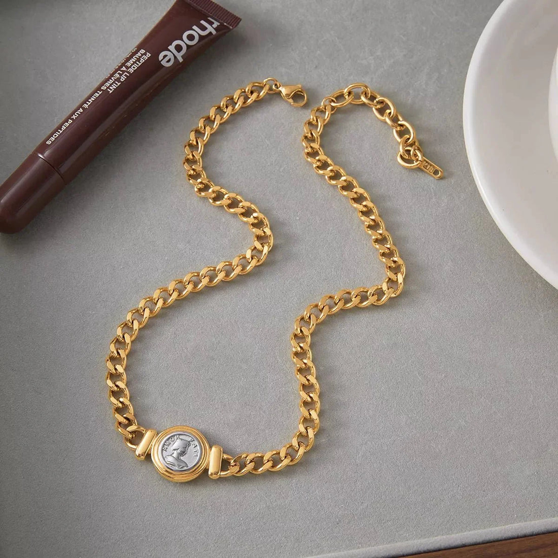 Gold chain necklace with a Roman coin pendant and a brown lip tint tube.