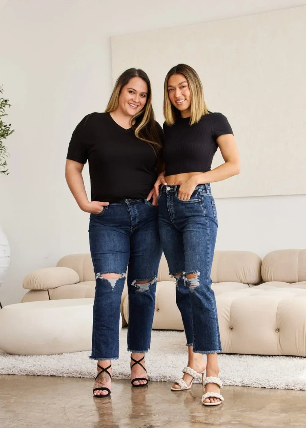 Two women in dark wash ripped jeans and black tops stand with hands in pockets.
