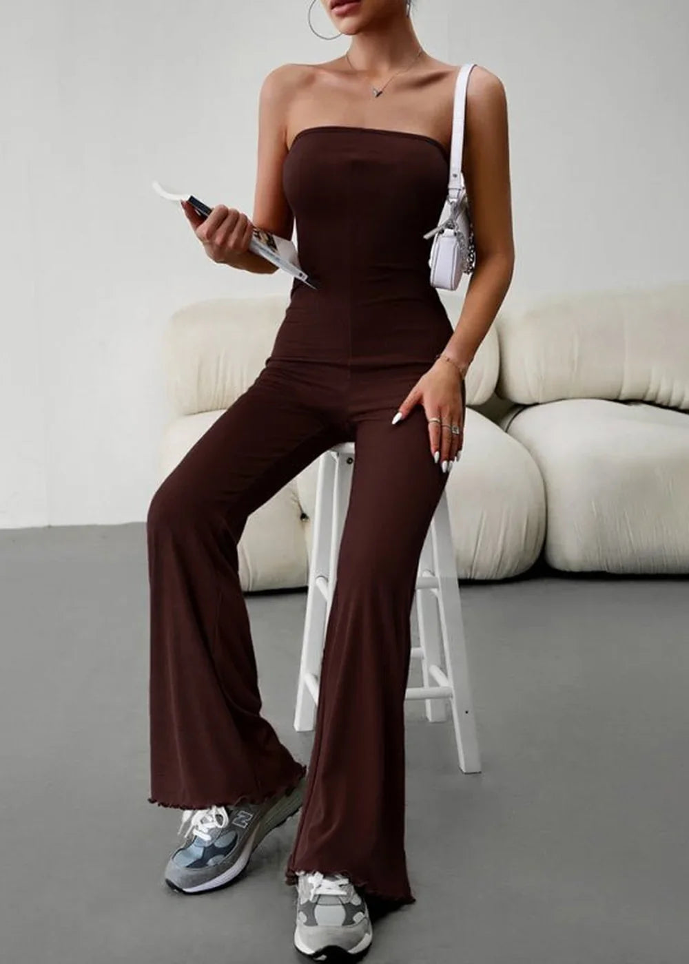 Brown strapless jumpsuit with flared legs and ruffled hem, worn by a model sitting on a stool.