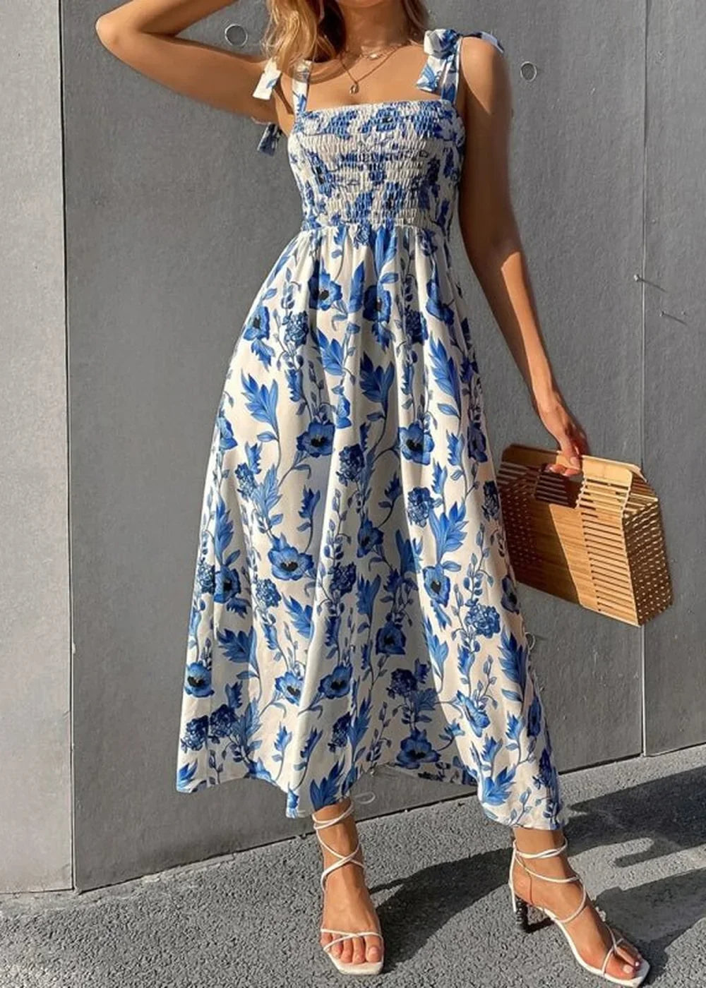 Blue and white floral print maxi dress with smocked bodice and tie straps, held by a wicker bag.