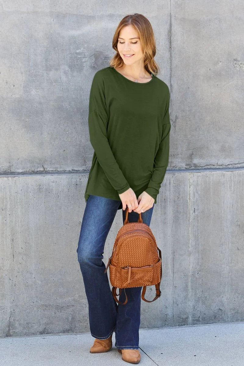 Woman wearing a long-sleeve olive green top and dark wash flare jeans, holding a woven brown backpack.