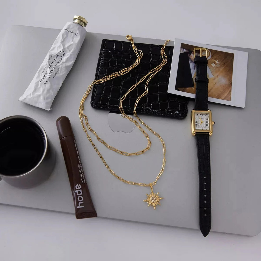 Gold layered necklace with sun pendant, black watch, and makeup on a laptop.