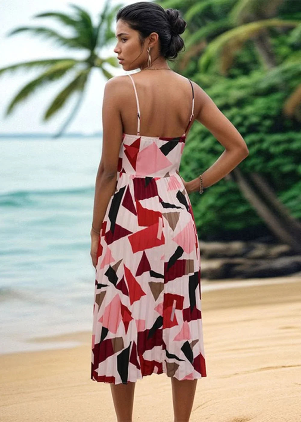 A woman in a pleated midi dress with a geometric print of red, pink, and black shapes stands on a beach.