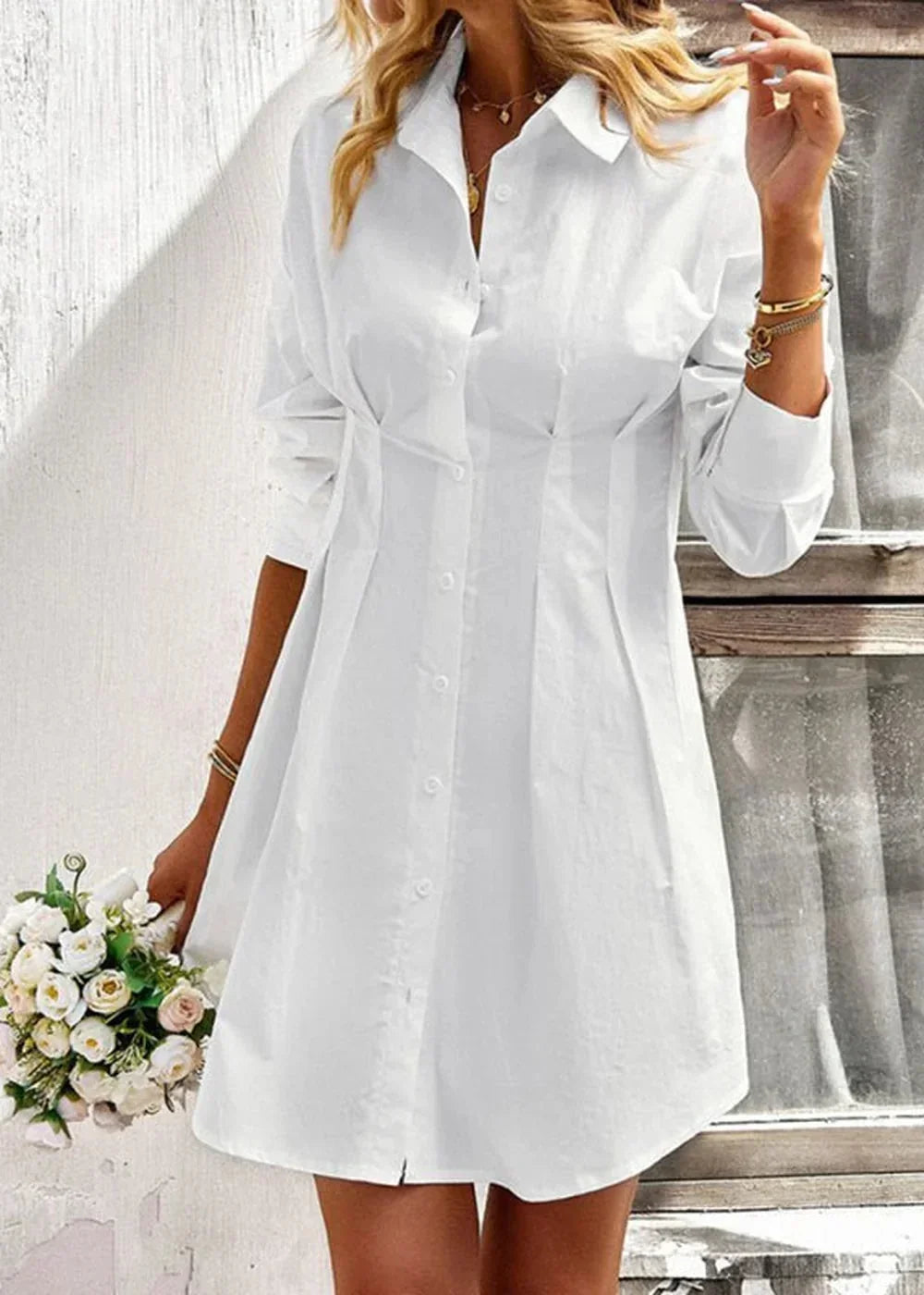 Crisp white cotton shirt dress with a cinched waist and button-front closure, held by a model with a bouquet of white flow...