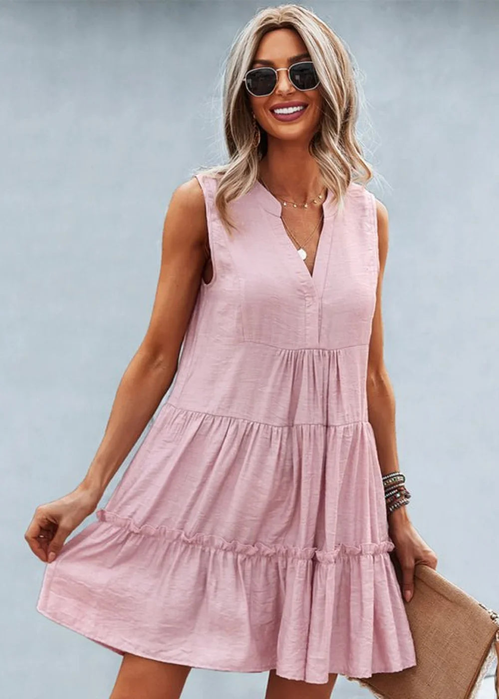 A woman in a pink sleeveless tiered dress with a v-neck and sunglasses smiles.