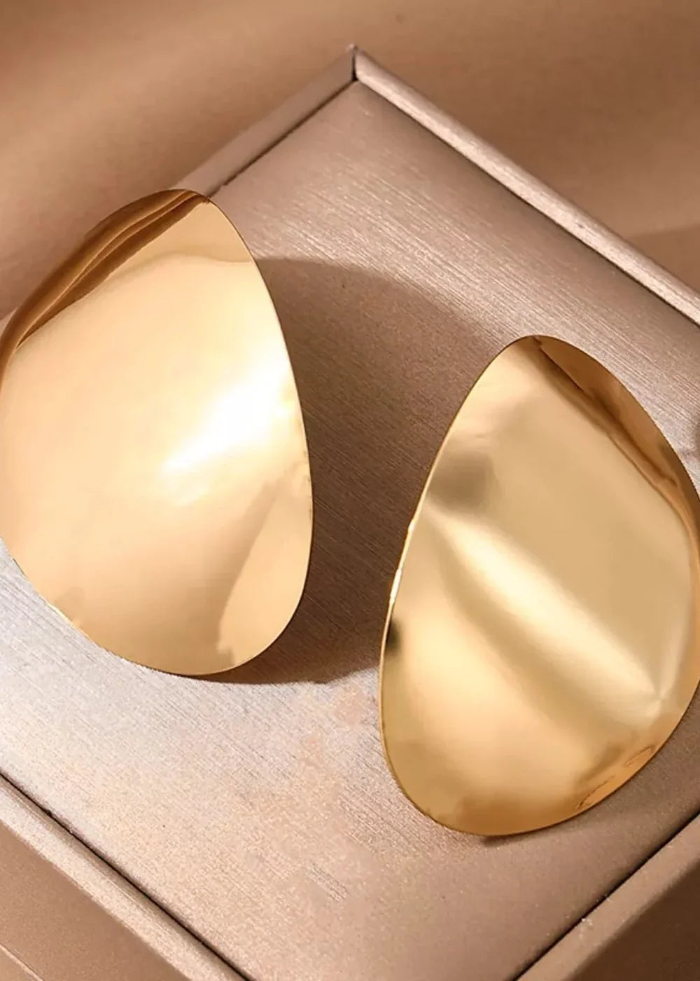 Golden, abstract, teardrop-shaped earrings with a high-shine, polished finish, resting on a textured beige surface.