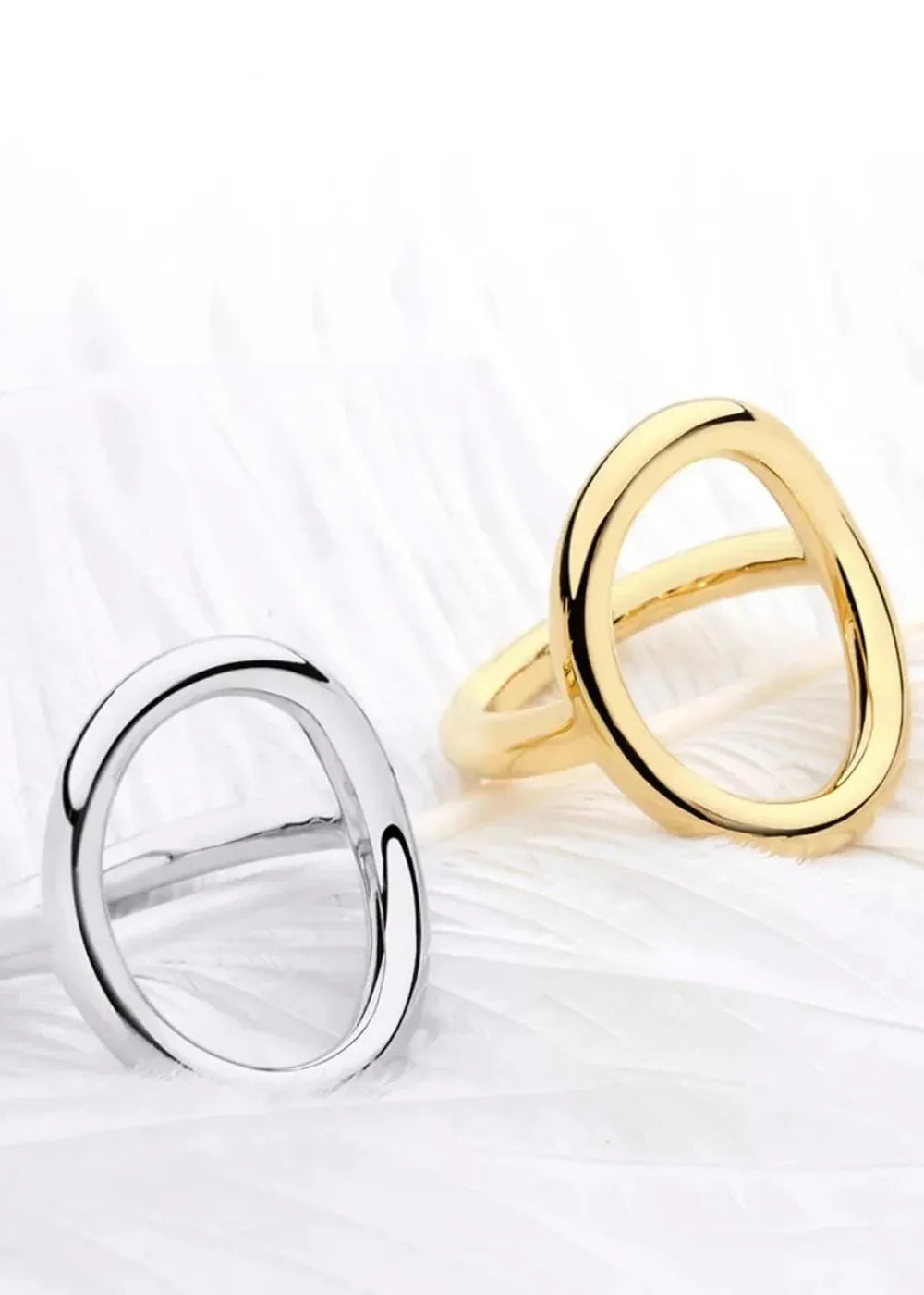 Two minimalist rings, one silver-plated and one 18K gold-plated, on a white feathery surface.