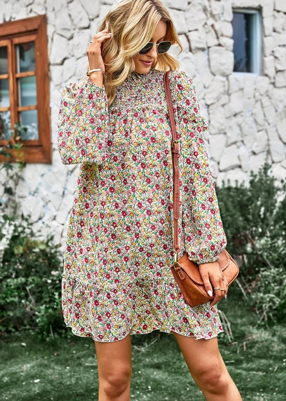 A woman models a floral print dress with a smocked neckline and ruffled hem, holding a brown crossbody bag.