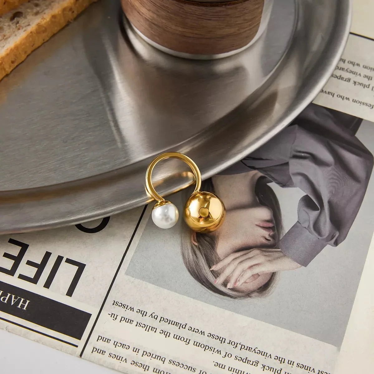 Gold ring with pearl and bead on a silver tray over a magazine page.