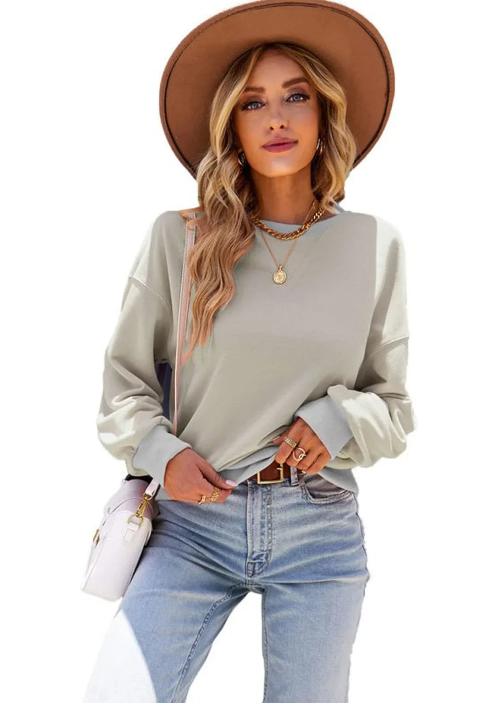 Woman in loose-fit light gray sweatshirt, blue jeans, and brown hat, wearing gold necklaces and rings.