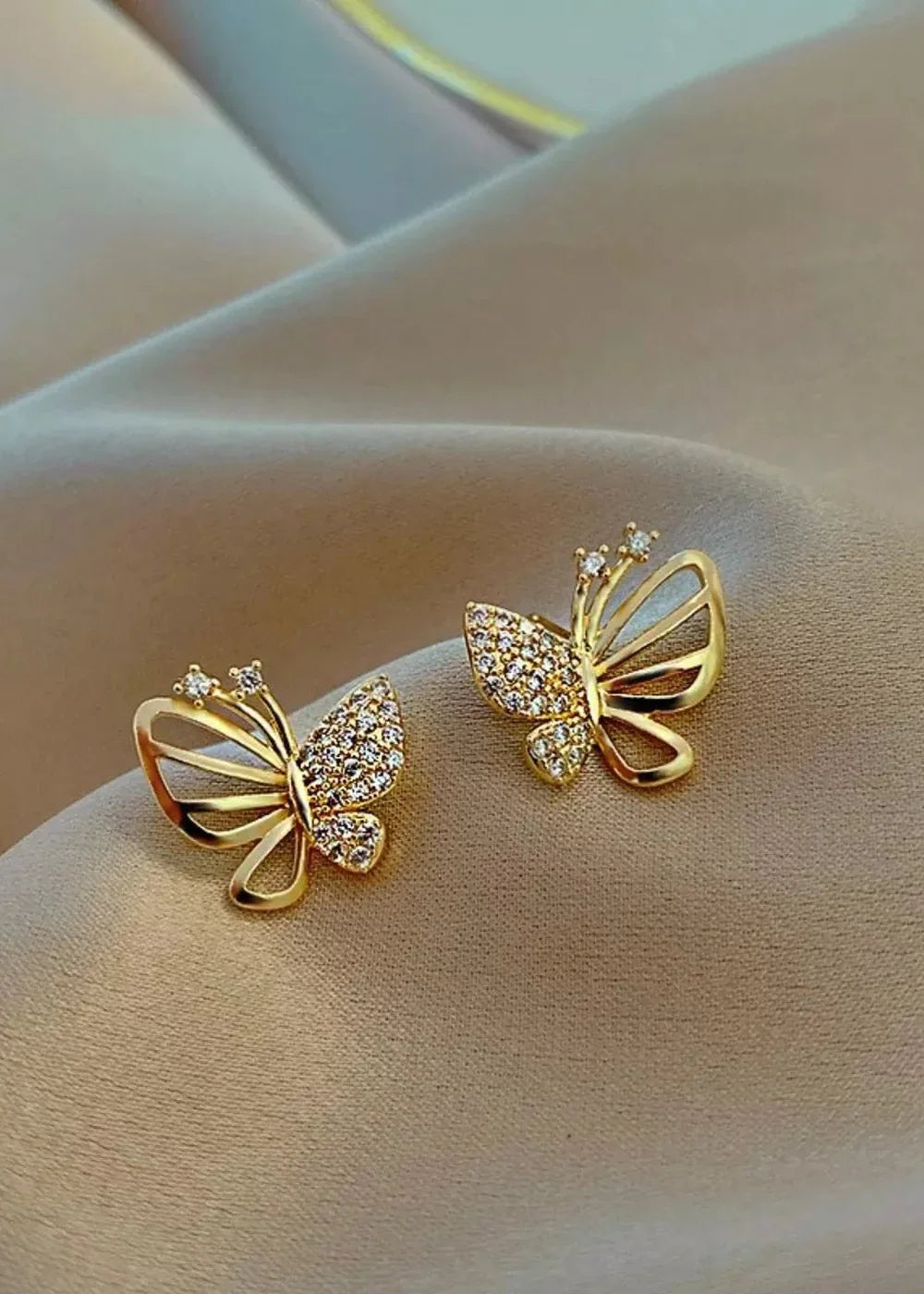 Gold butterfly earrings with rhinestones and open wings, resting on a tan fabric.