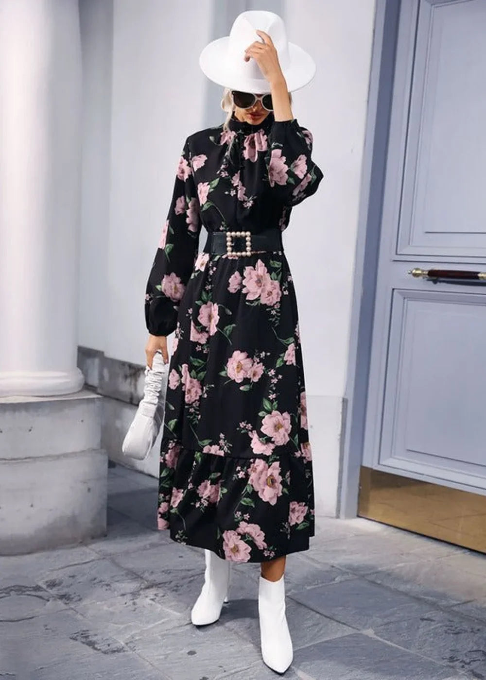 Woman in a black floral print midi dress, white hat, sunglasses, and white boots.