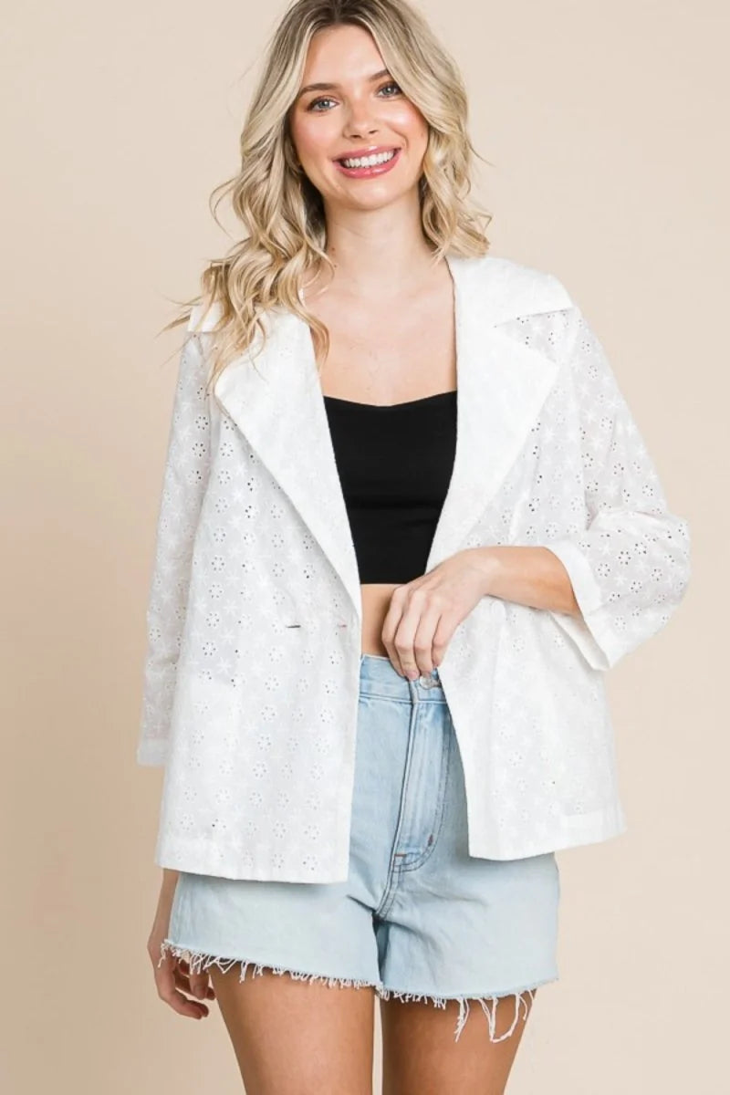 White eyelet jacket with wide lapels and three-quarter sleeves, paired with light wash denim shorts.