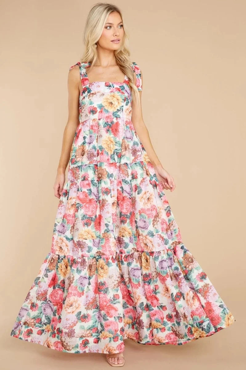 A woman models a flowing tiered maxi dress with a vibrant floral pattern in pink, yellow, and green on a light background.